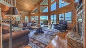 A spacious log cabin interior with a large living area, multiple seating options, and large windows offering views of the outdoors.
