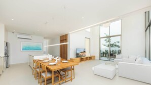 A modern living room with a white sofa, a dining area with a wooden table and chairs, and a large window offering a view of the outdoors.