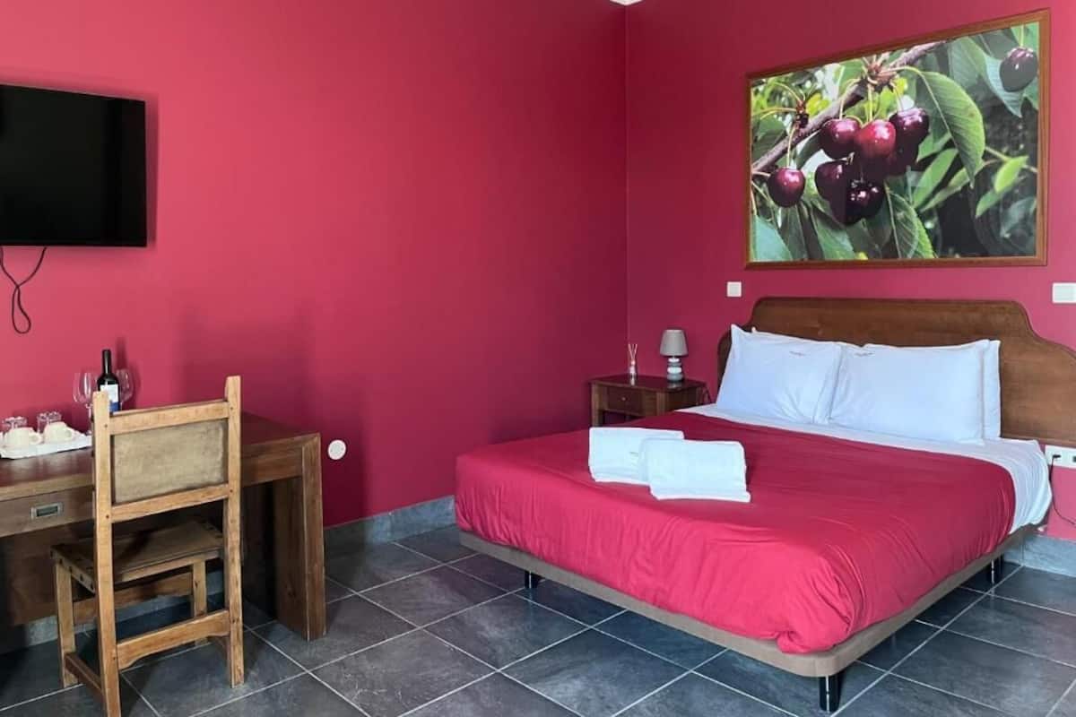A hotel room with a bed, a desk, a chair, a TV, and a painting of cherries on the wall.