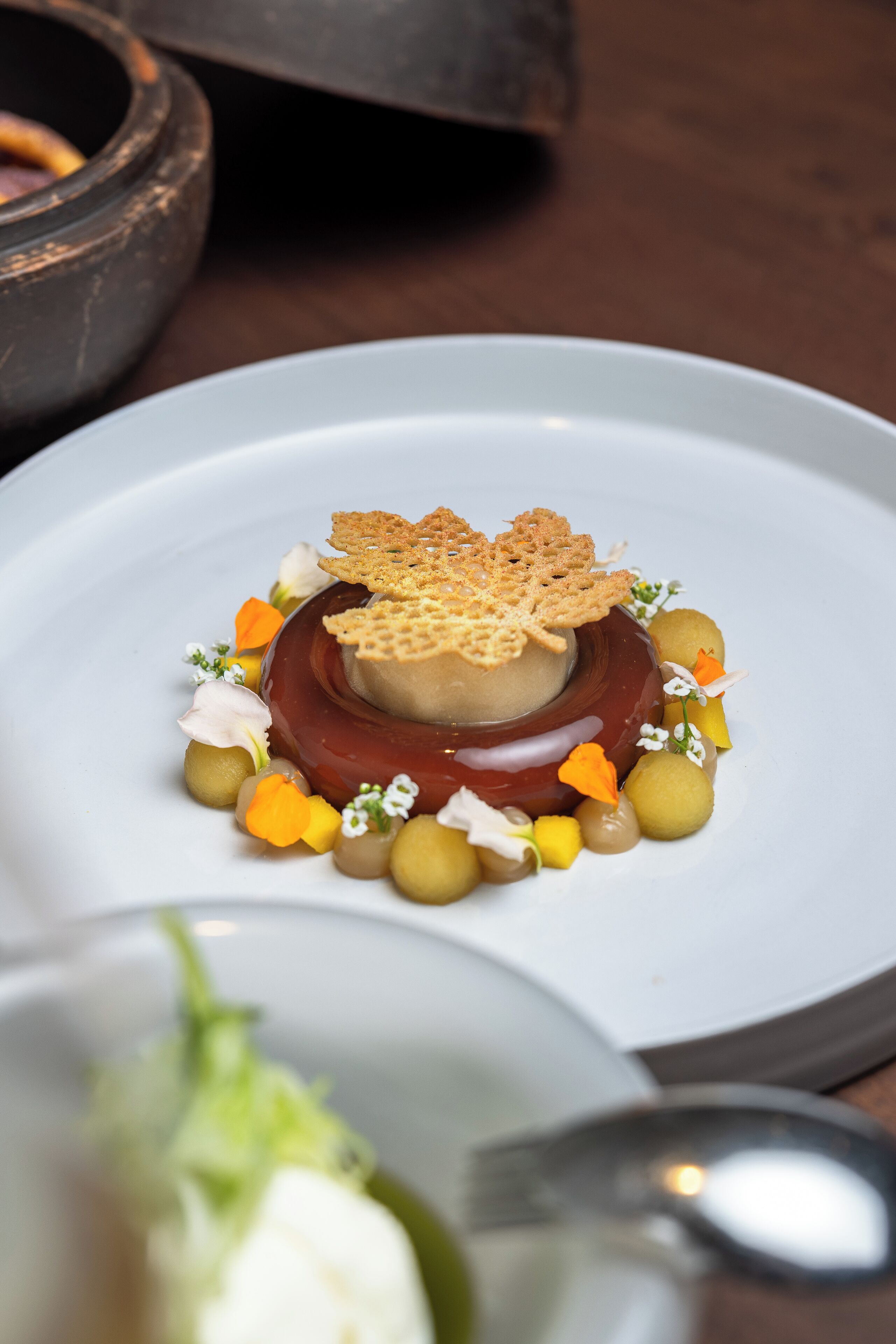 A dessert on a white plate with a central spherical dessert, surrounded by small round items and garnished with flowers and leaves.
