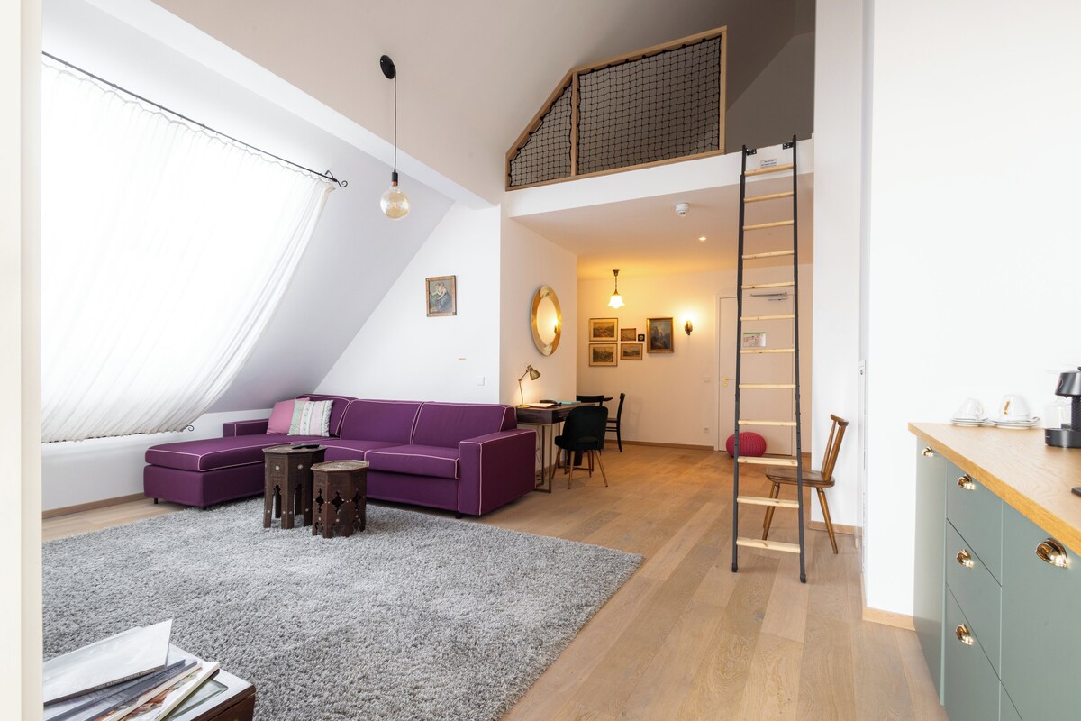 Un dormitorio estilo loft con techo inclinado, un sofá morado, una escalera de madera y una zona de escritorio.