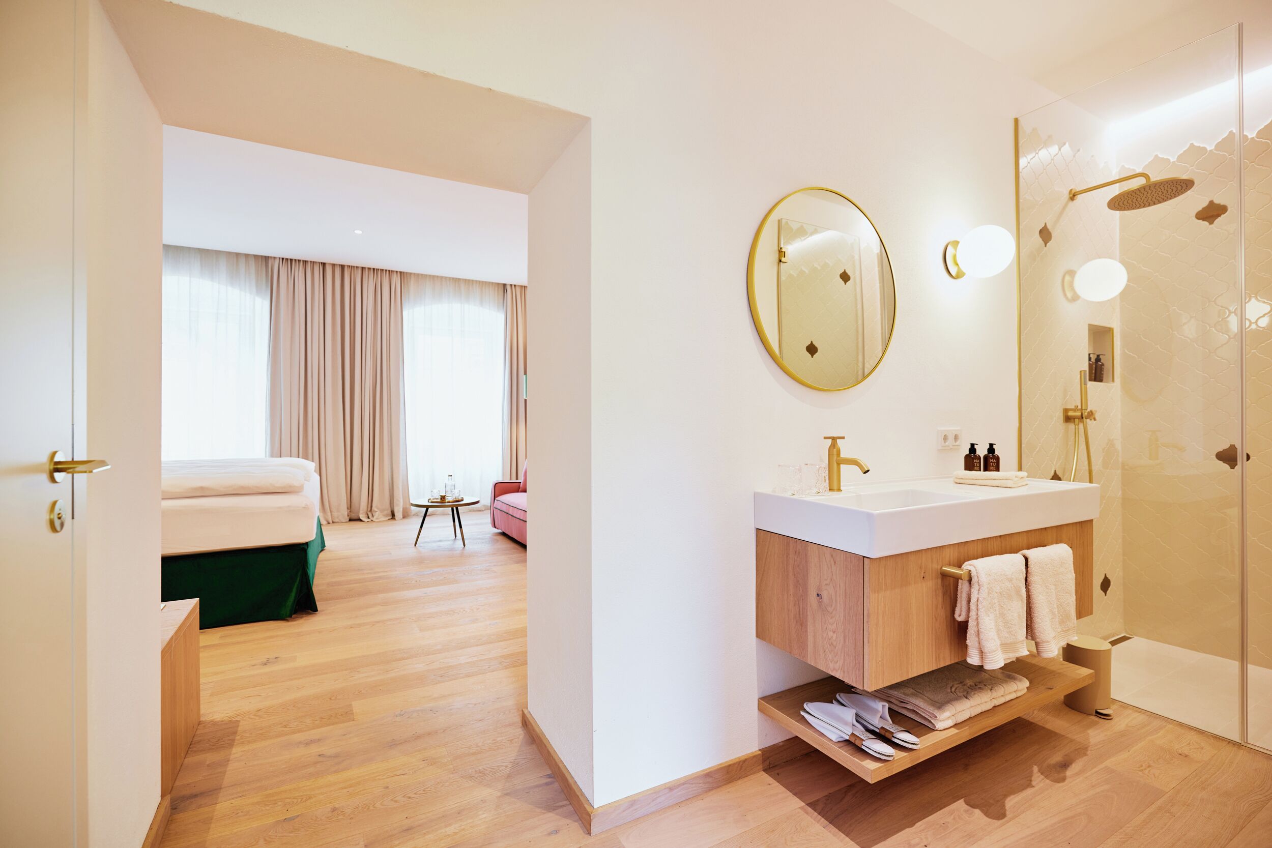 Une chambre d’hôtel moderne avec un plancher en bois, un grand lit, une salle de bain dotée d’un lavabo et d’un miroir blancs, et un espace douche.
