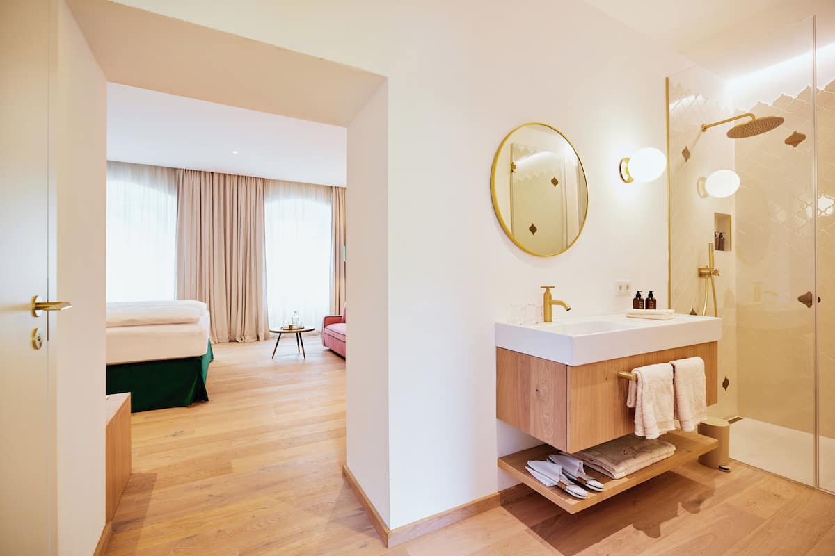 Une chambre d’hôtel moderne avec un plancher en bois, un grand lit, une salle de bain dotée d’un lavabo et d’un miroir blancs, et un espace douche.