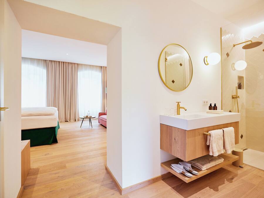 A modern hotel room with a wooden floor, a large bed, a bathroom with a white sink and mirror, and a shower area.