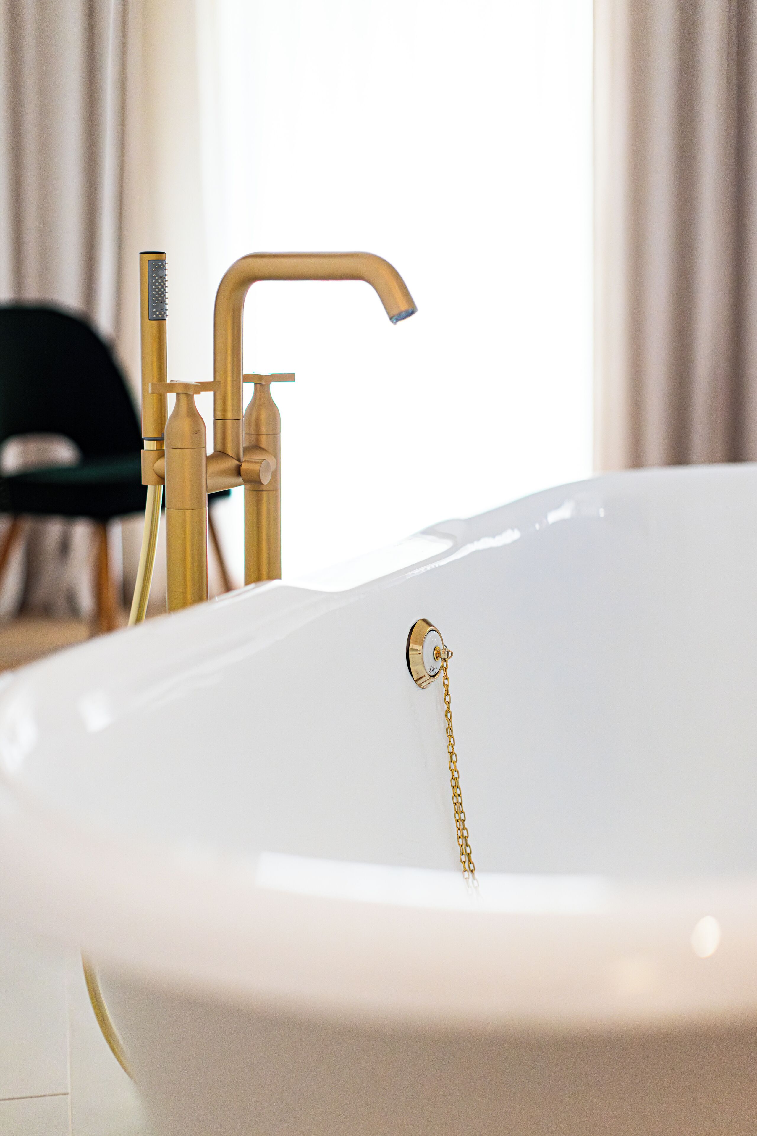 Una bañera blanca con grifo y ducha dorados, una cadena dorada y una silla verde al fondo.