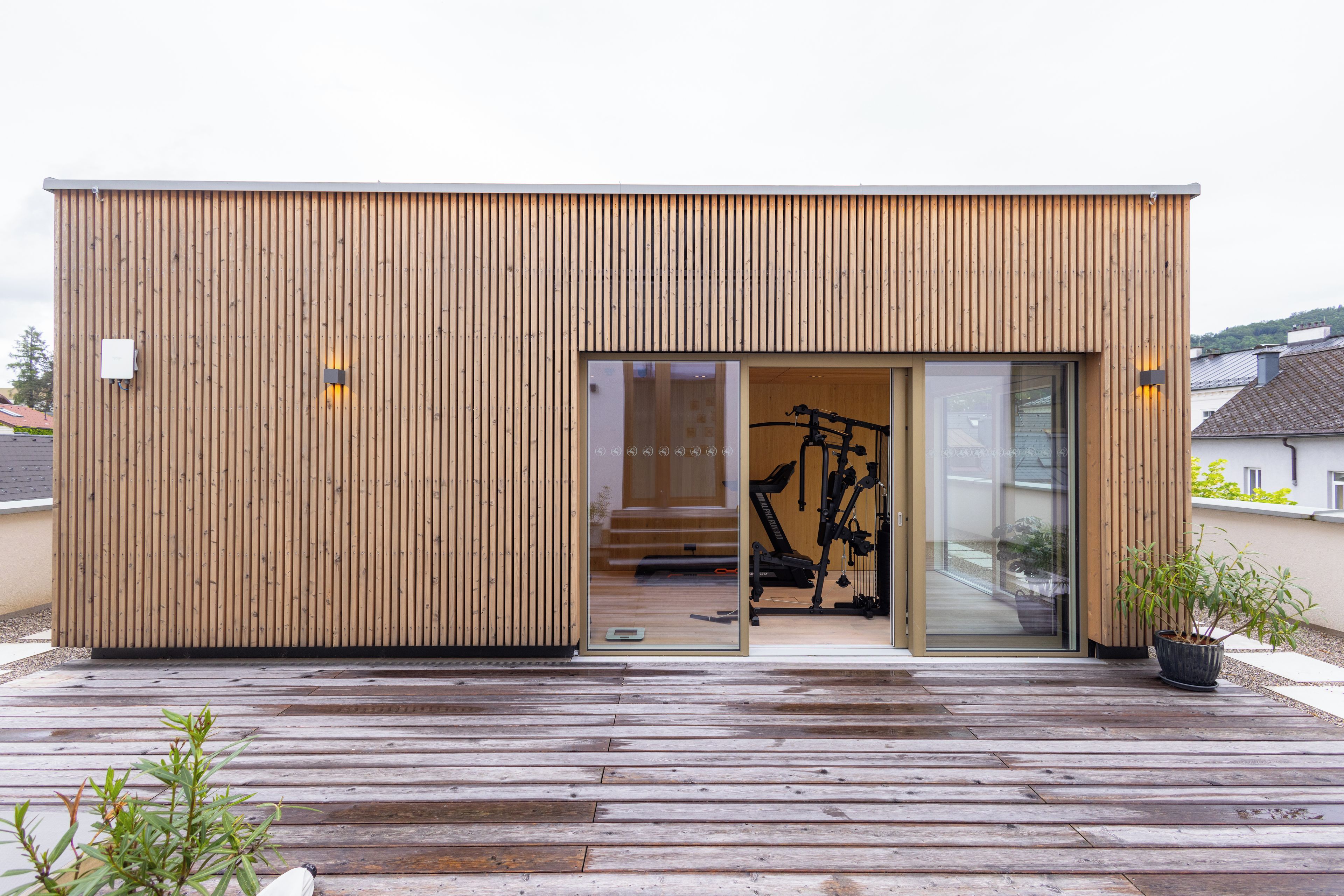 A modern wooden building with a sliding glass door entrance, a visible exercise bike inside, and a wooden deck outside.