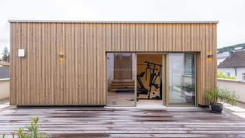 Un edificio moderno de madera con puerta de vidrio corrediza, una bicicleta estática visible en su interior y una terraza de madera en el exterior.
