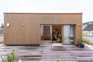 A modern wooden building with a sliding glass door entrance, a visible exercise bike inside, and a wooden deck outside. - Boutique Hotel Zum Goldenen Hirschen (Gmunden)