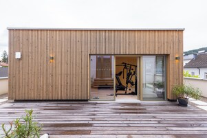 Un edificio moderno de madera con puerta de vidrio corrediza, una bicicleta estática visible en su interior y una terraza de madera en el exterior.