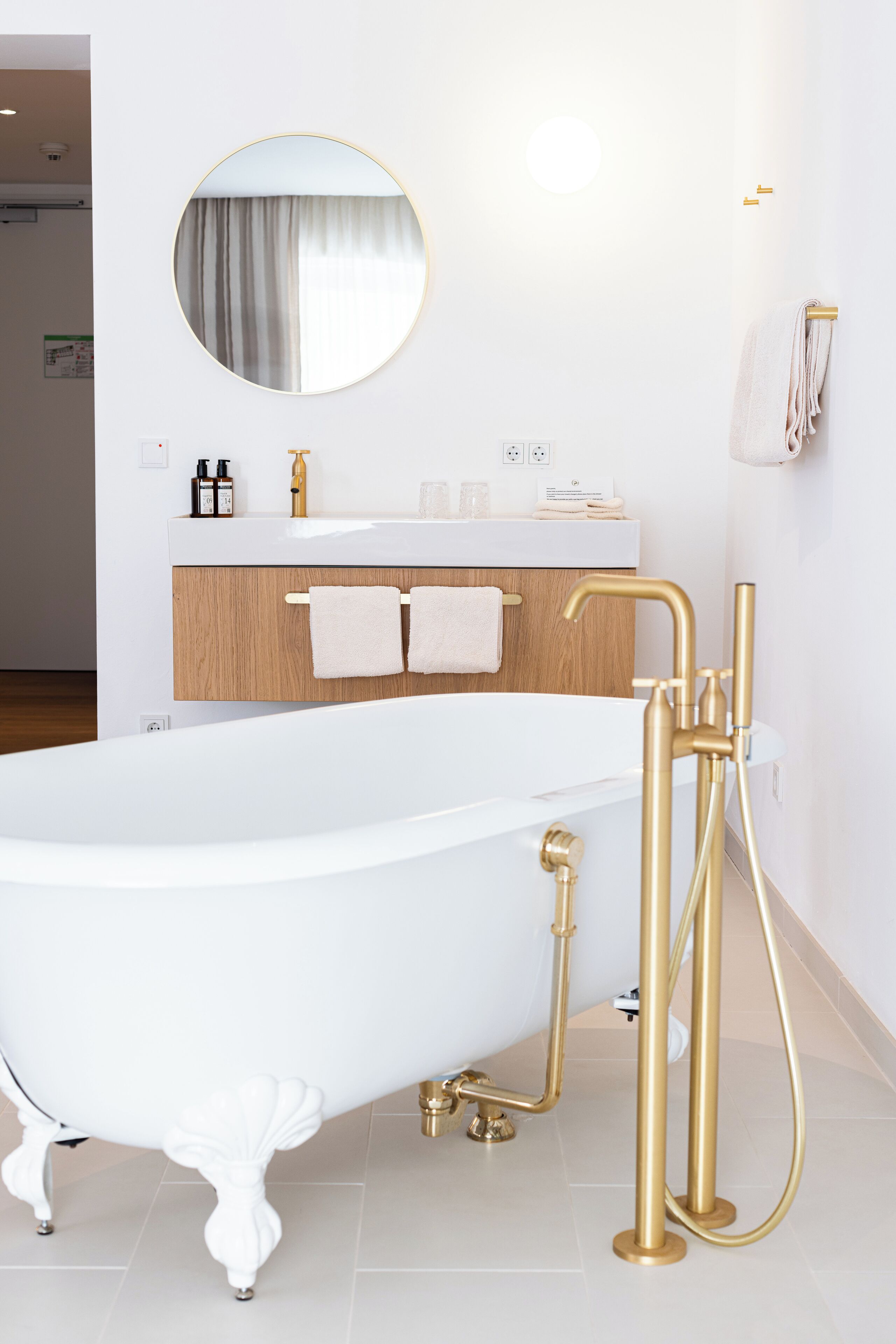 Een moderne badkamer met een vrijstaand bad, een houten wastafelmeubel, een ronde spiegel en gouden details.