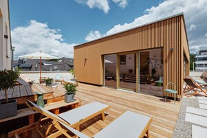 Una terraza moderna al aire libre con mobiliario de madera y un ventanal de vidrio que da a un espacio interior.