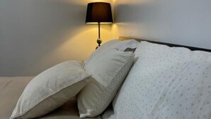 A bed with two pillows and a blanket, a wall-mounted lamp, and a wooden headboard.