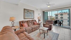 A spacious living room with a large sofa, a coffee table, and a chair. There is a painting on the wall and a window with a view of buildings.