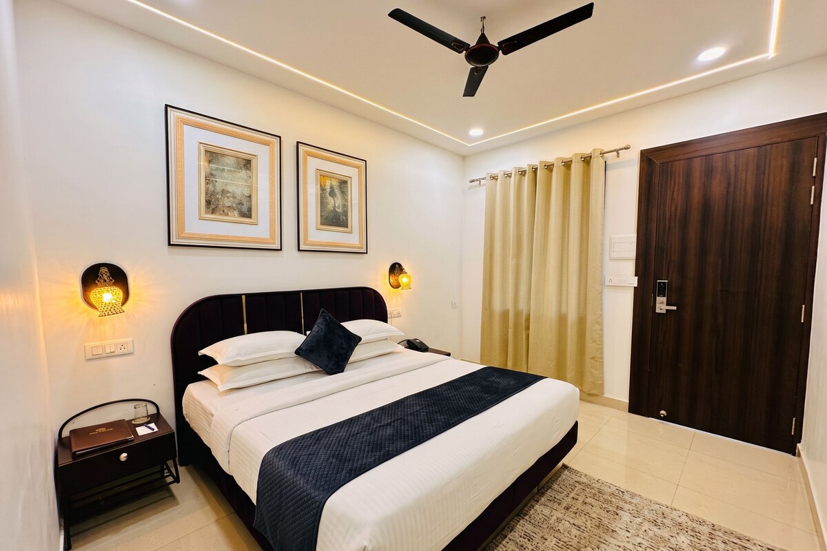Une chambre à coucher avec un lit, des tables de chevet, un ventilateur de plafond, des œuvres d’art encadrées et une porte.