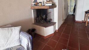 Une pièce chaleureuse avec une cheminée, une étagère en bois remplie de livres et de bocaux, un coussin à motifs et un sol carrelé.