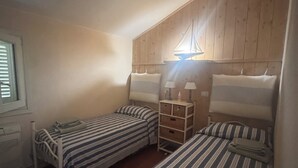 Une petite chambre agréable avec deux lits simples, une tête de lit en bois, une table de chevet avec une lampe et une fenêtre avec des stores.