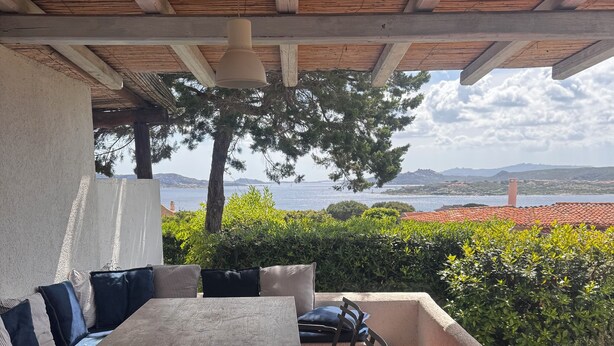 Une terrasse extérieure avec une table et des chaises en bois, un canapé avec des coussins, et une vue sur la mer et les collines en arrière-plan.