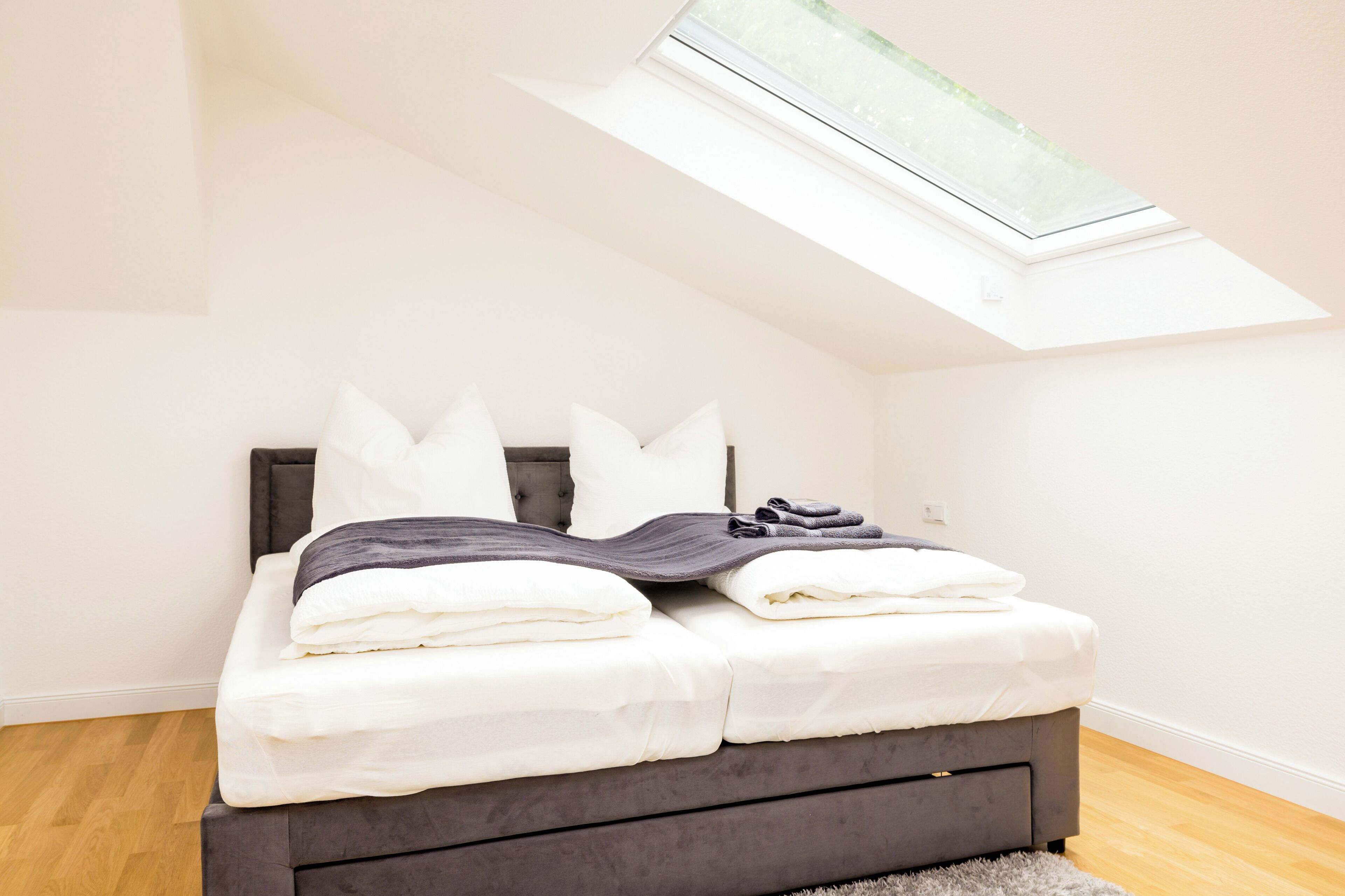 Une chambre avec une lucarne, un lit recouvert de draps blancs et une tête de lit grise.