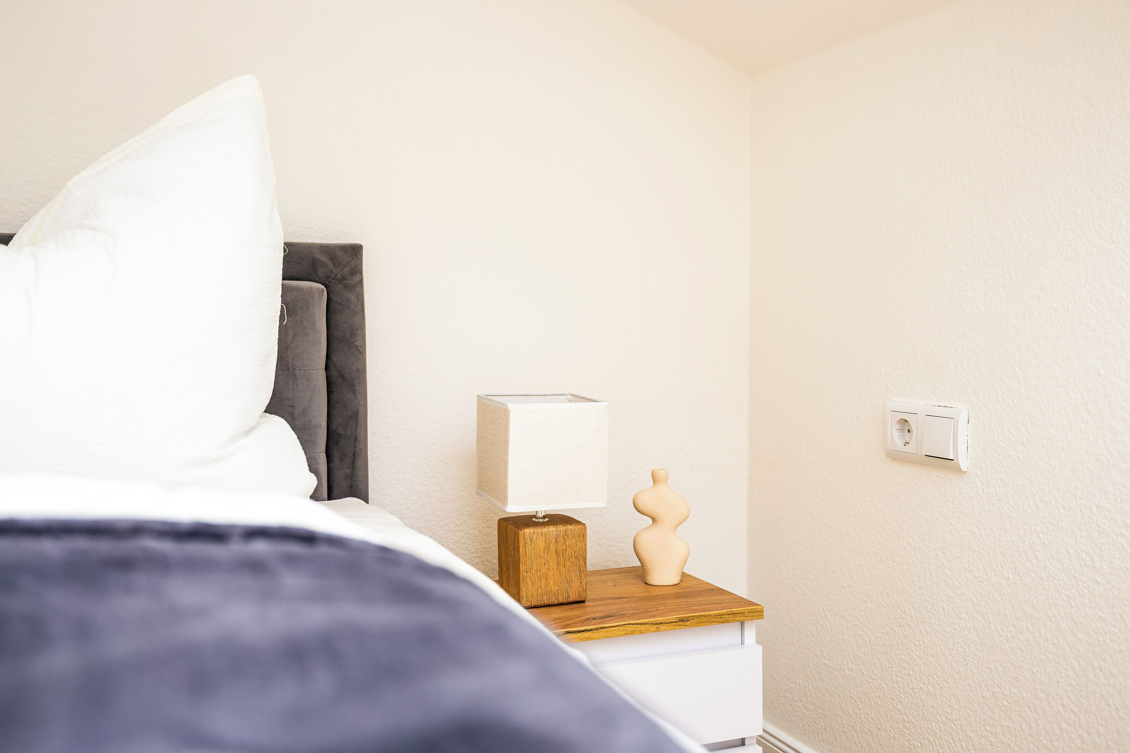 Cama bem arrumada com roupa de cama branca e manta azul escura, mesa de cabeceira de madeira com abajur branco e um objeto decorativo, e parede lisa com interruptor de luz.