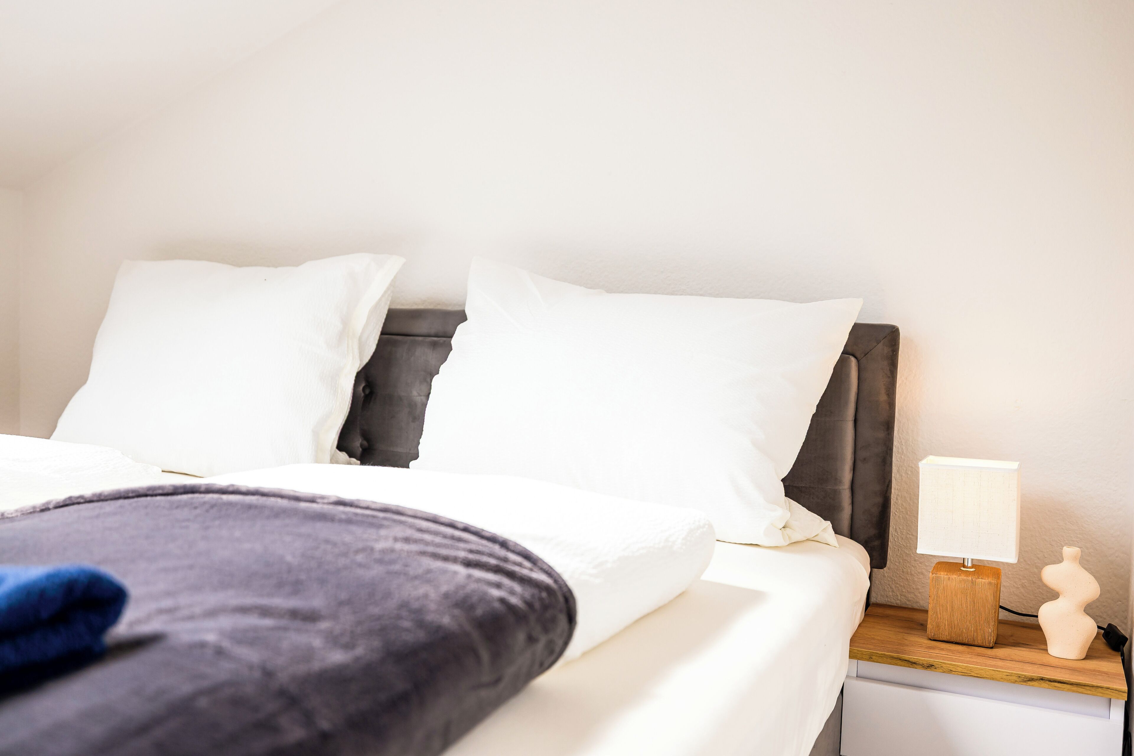 Una cama bien hecha con sábanas blancas y una manta gris oscuro, una mesita de noche de madera con una lámpara y un pequeño jarrón.
