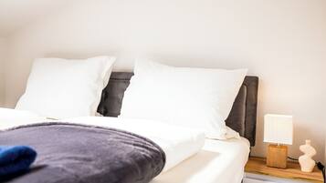 Una cama bien hecha con sábanas blancas y una manta gris oscuro, una mesita de noche de madera con una lámpara y un pequeño jarrón.
