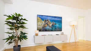 A modern living room with a large painting of a coastal landscape, a potted plant, a white console table, and a floor lamp. - NORA Dresden Hauptbahnhof (Dresden)