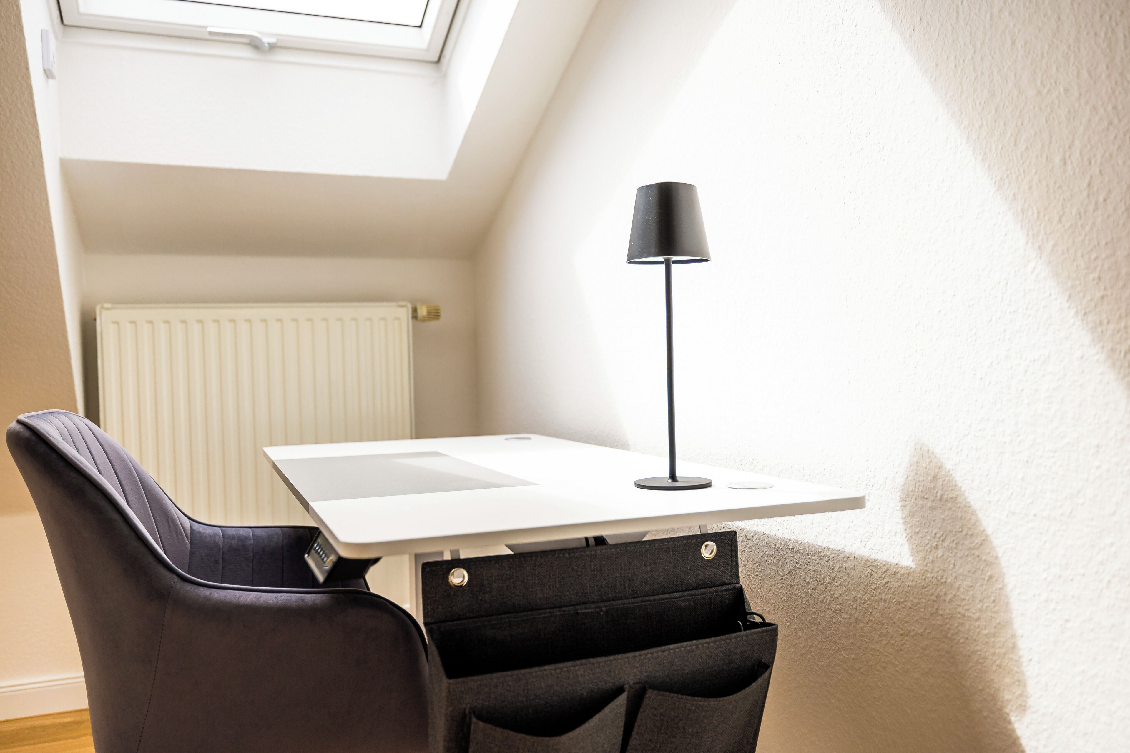 A small, well-lit room with a white desk, a black chair, a black floor lamp, and a radiator.