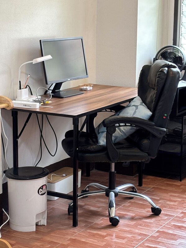 A workspace with a wooden desk, a black office chair, a computer, a desk lamp, a white trash bin, and a standing fan.