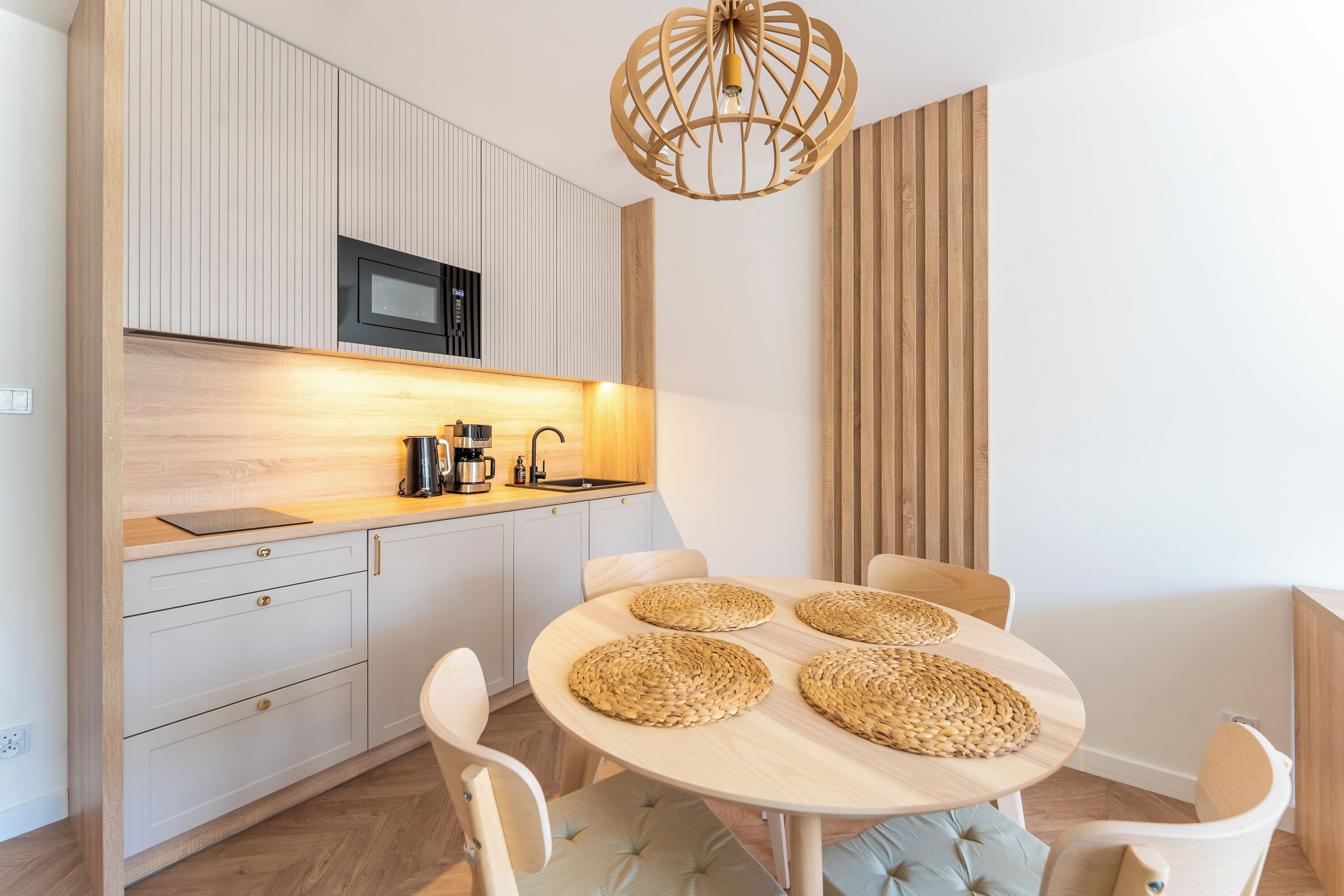 Une cuisine moderne équipée d’un four à micro-ondes, d’une machine à café et de meubles en bois. On y trouve une table ronde avec des sets de table en fibres tressées et des chaises rembourrées.