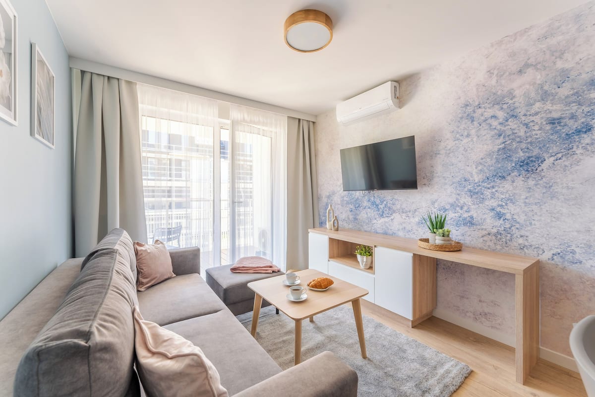 Un salon moderne comprenant un canapé, une table basse, un téléviseur fixé sur un mur à texture, et un petit meuble agrémenté d’objets décoratifs.