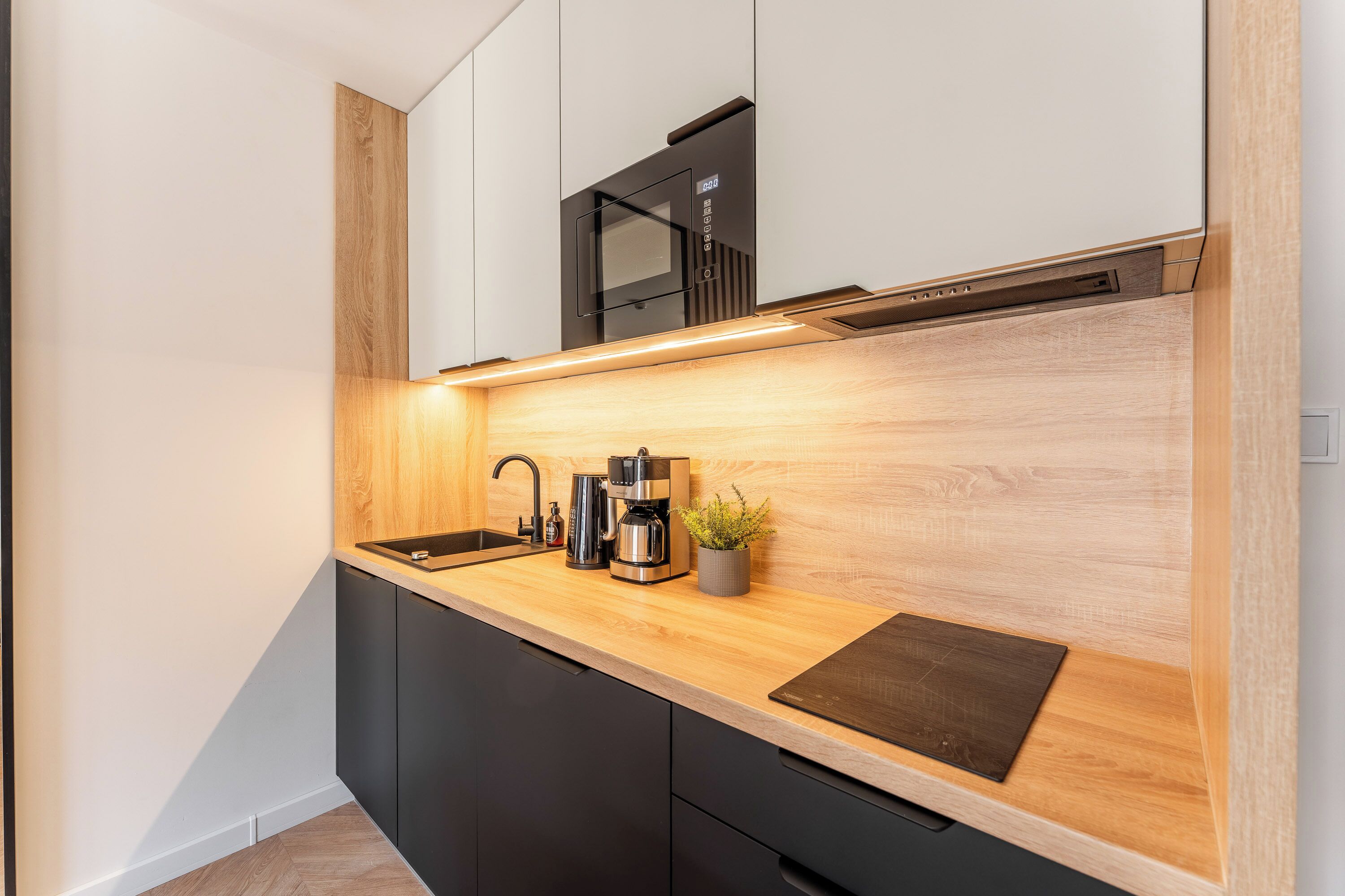 A modern kitchen with a wooden countertop, black cabinets, a built-in microwave, a coffee maker, and a small potted plant.