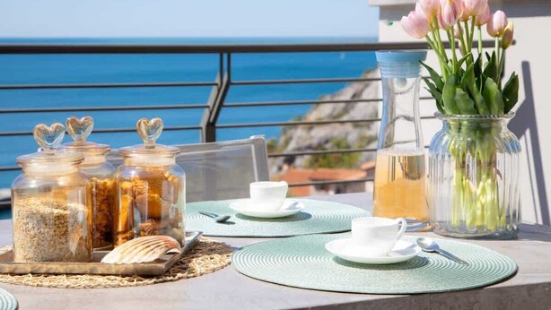 Una mesa dispuesta con frascos de vidrio, un jarrón con flores y una jarra de cristal, con vista a un cuerpo de agua.