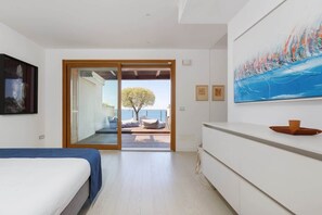 A modern room with a bed, a white dresser, a large abstract painting, and a sliding glass door leading to an outdoor seating area with a tree and a view of the sea.