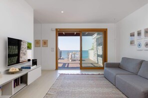 A modern living room with a flat-screen TV, a grey sofa, a patterned rug, and a sliding glass door leading to a balcony with a view of the sea.