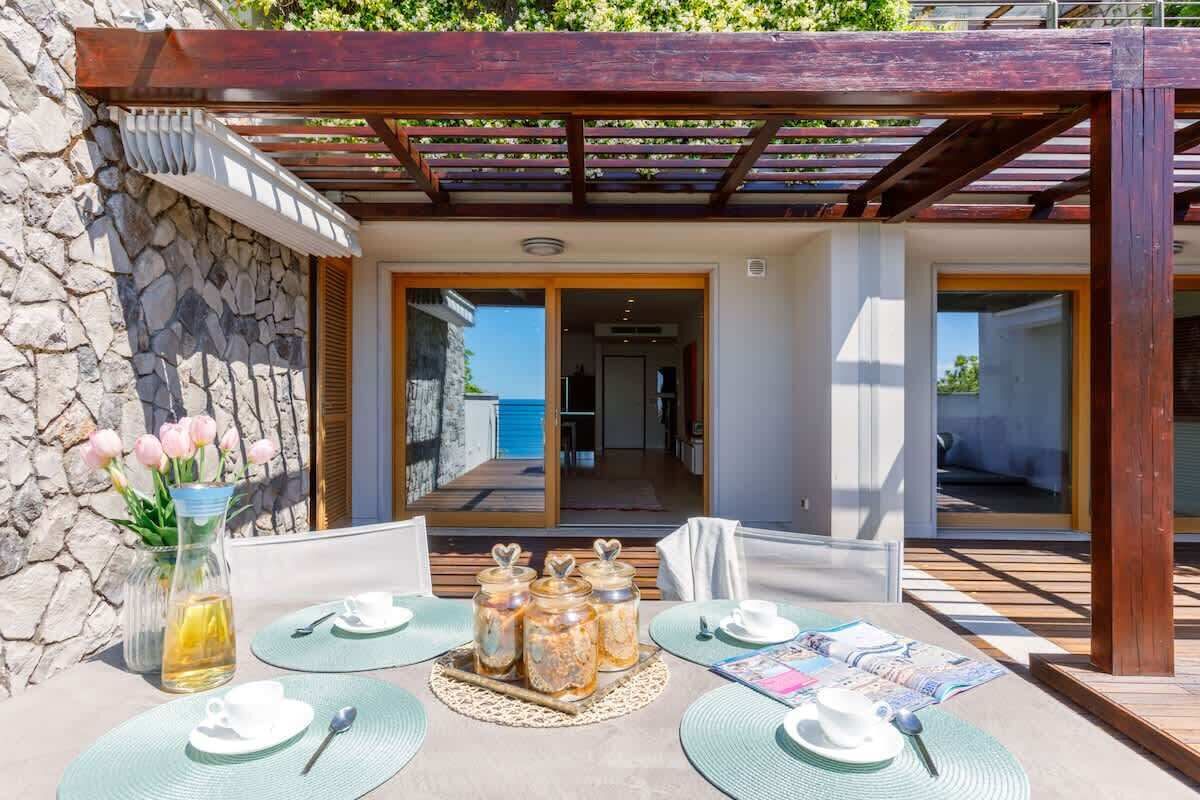 A covered outdoor dining area with a stone wall, a wooden pergola, and a view into the interior.