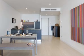 A modern kitchen with a dining area, a small table with a magazine, and a colorful abstract painting on the wall. - Ulivo sul golfo - Portopiccolo Apartments (Duino-Aurisina)