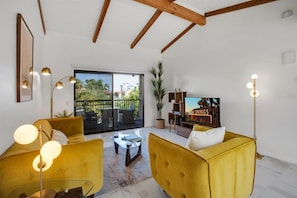 Sala de estar com sofás amarelos, mesa de centro de vidro, televisão e varanda com vista.