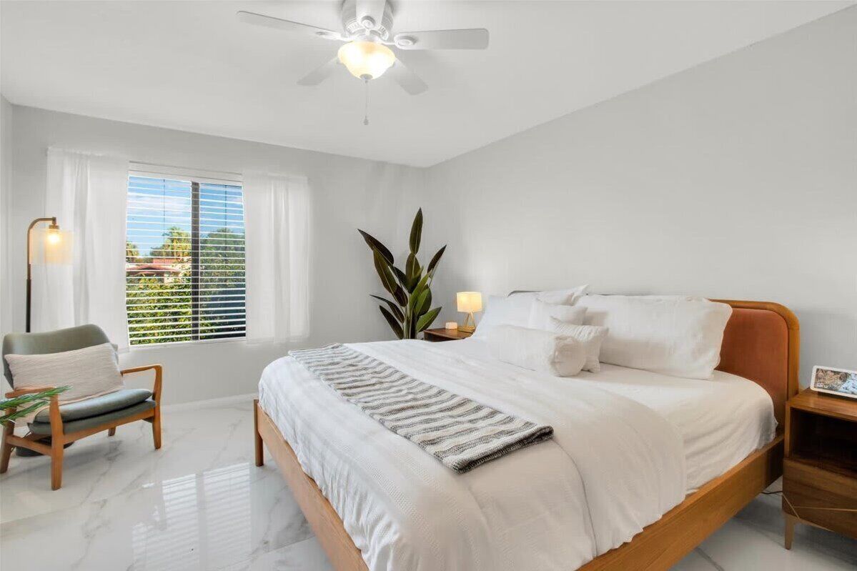 A bedroom with a bed, a chair, a nightstand, a plant, and a ceiling fan.