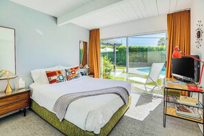 A bedroom with a bed, a desk with a TV, and a view of a pool and greenery.
