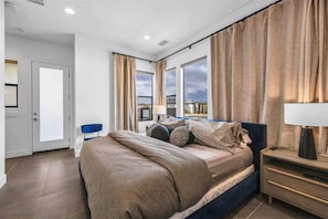 A bedroom with a bed, a nightstand, a chair, and a view of the outside.