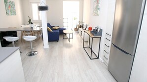 A modern kitchen with a stainless steel refrigerator, a dining area with a white table and chairs, and a blue sofa with cushions.