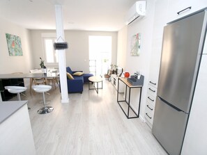 Une cuisine moderne avec un réfrigérateur en acier inoxydable, un coin repas avec une table et des chaises blanches, et un canapé bleu agrémenté de coussins.