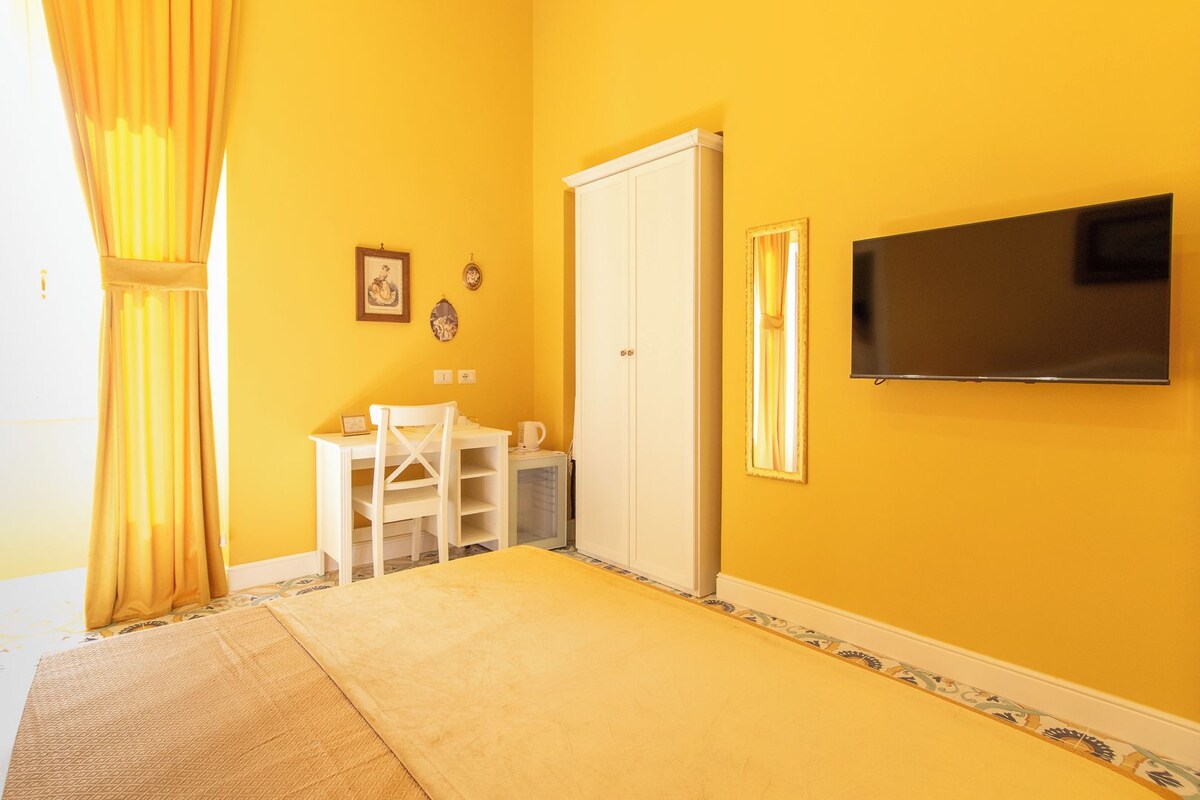 A room with yellow walls, a white door, a TV mounted on the wall, a small table with a chair, and a window with curtains.