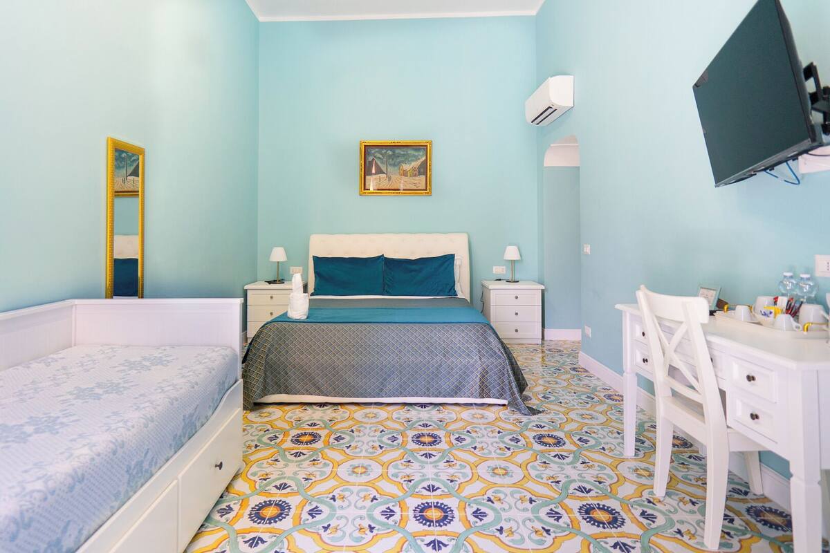 A bedroom with a bed, a desk, a chair, a TV, and a painting on the wall.