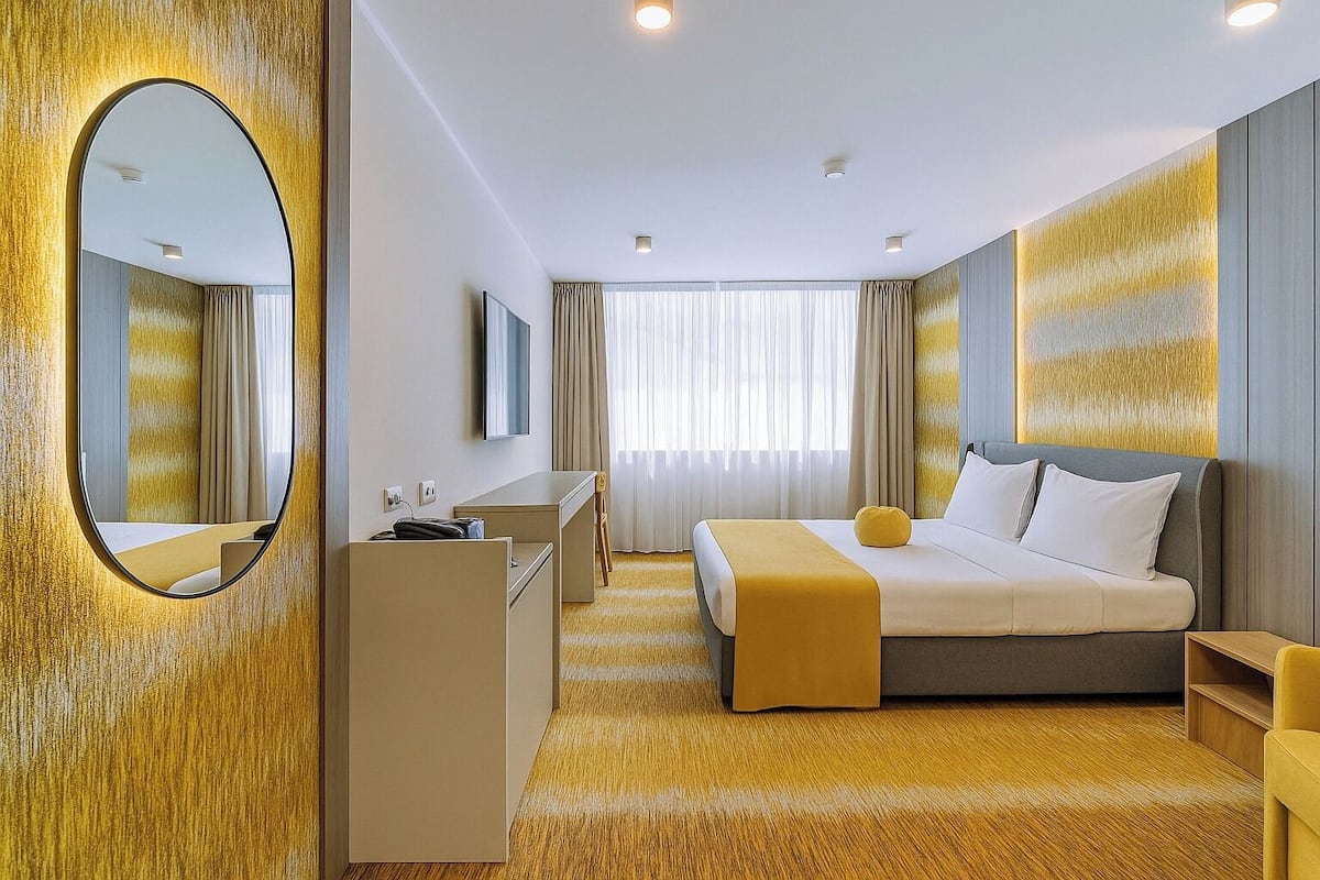 A modern hotel room with a large bed, a desk, a chair, and a mirror.