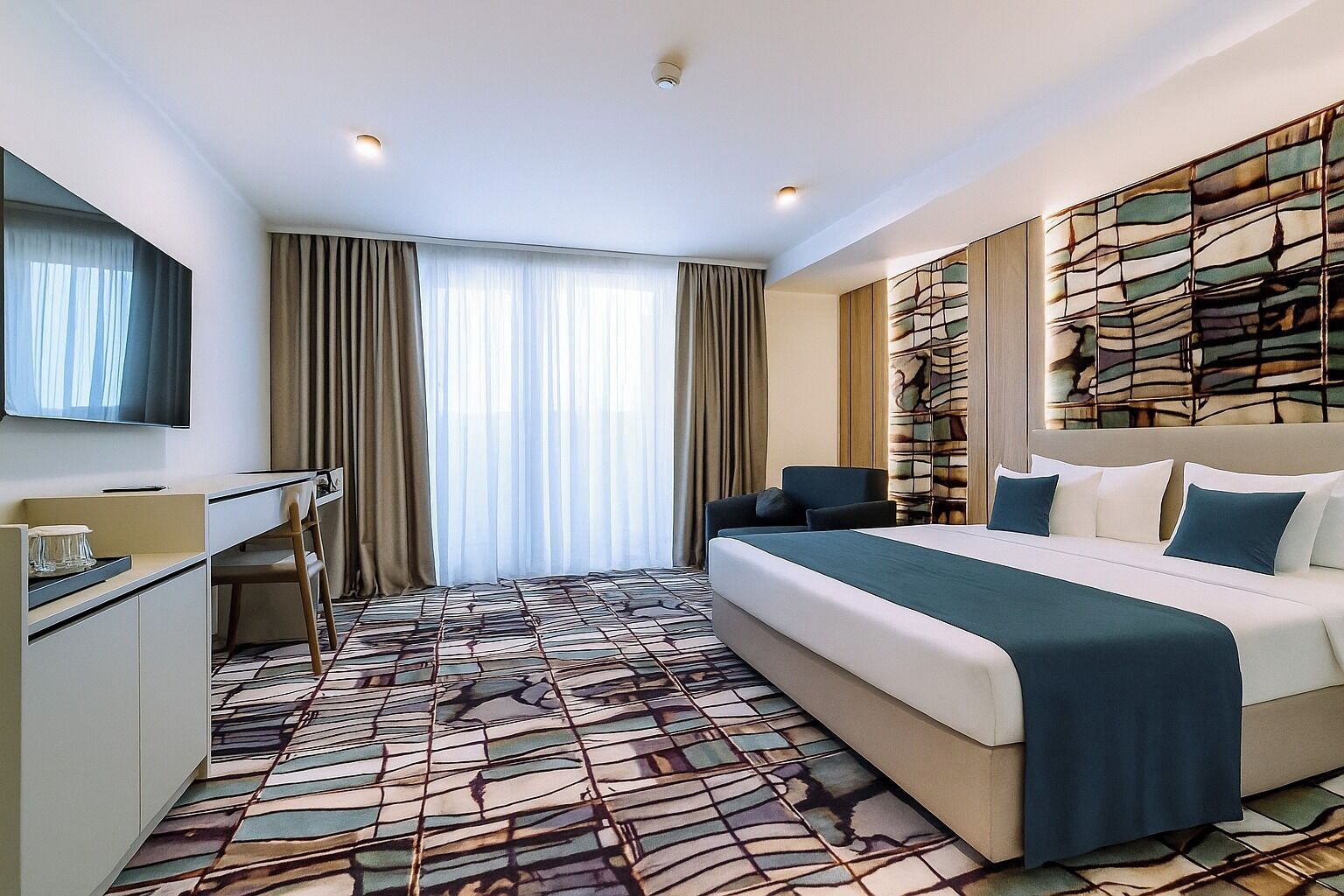 A modern hotel room with a large bed, a desk, a chair, and a TV.