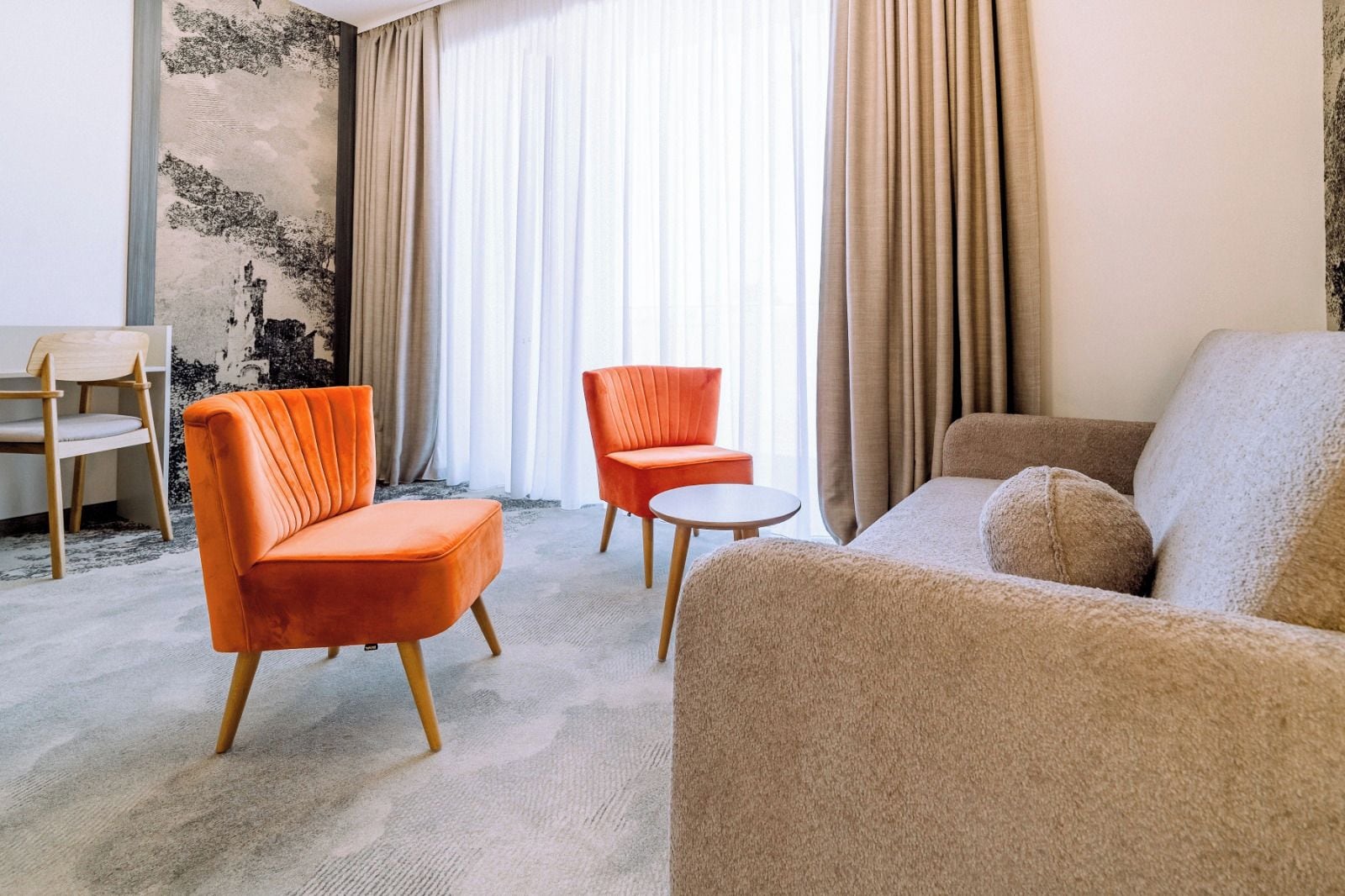 Ein modernes Wohnzimmer mit einer Couch, zwei Sesseln, einem kleinen runden Tisch und einem Sessel. Das Zimmer verfügt über ein großes Fenster mit Vorhängen, eine strukturierte Wand und einen Teppichboden.