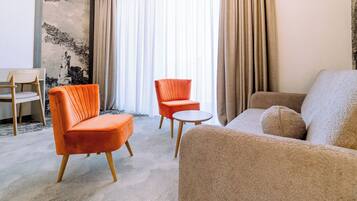 Ein modernes Wohnzimmer mit einer Couch, zwei Sesseln, einem kleinen runden Tisch und einem Sessel. Das Zimmer verfügt über ein großes Fenster mit Vorhängen, eine strukturierte Wand und einen Teppichboden.