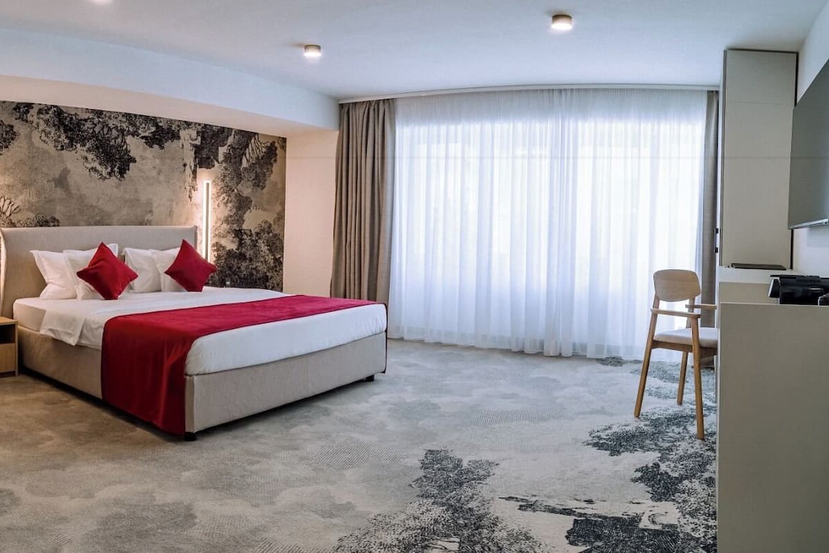 A modern hotel room with a large bed, a red armchair, a desk, and a large window with sheer curtains.