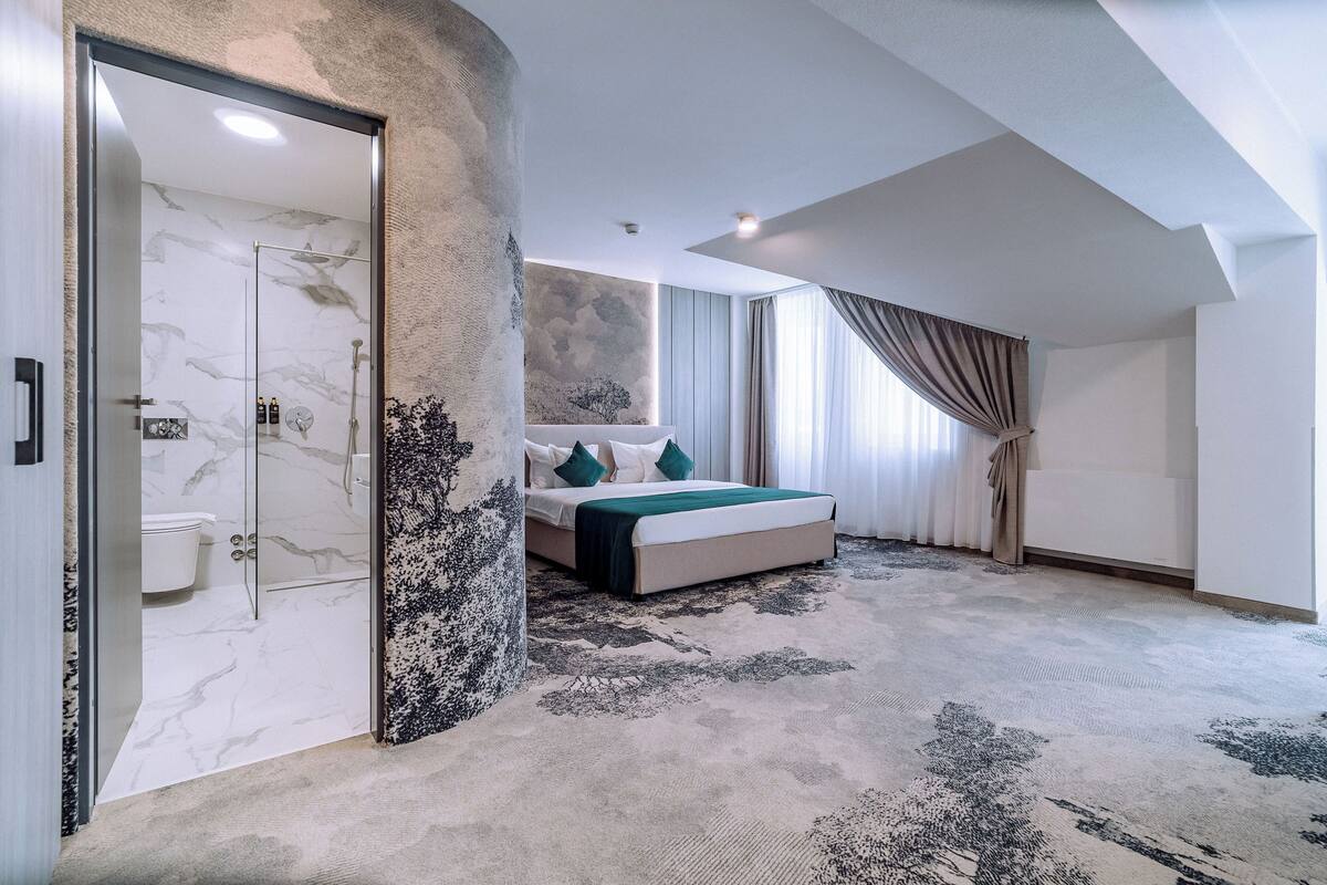 A modern bedroom with a marble shower, a large bed with a green throw, and a view of the outdoors through a large window.